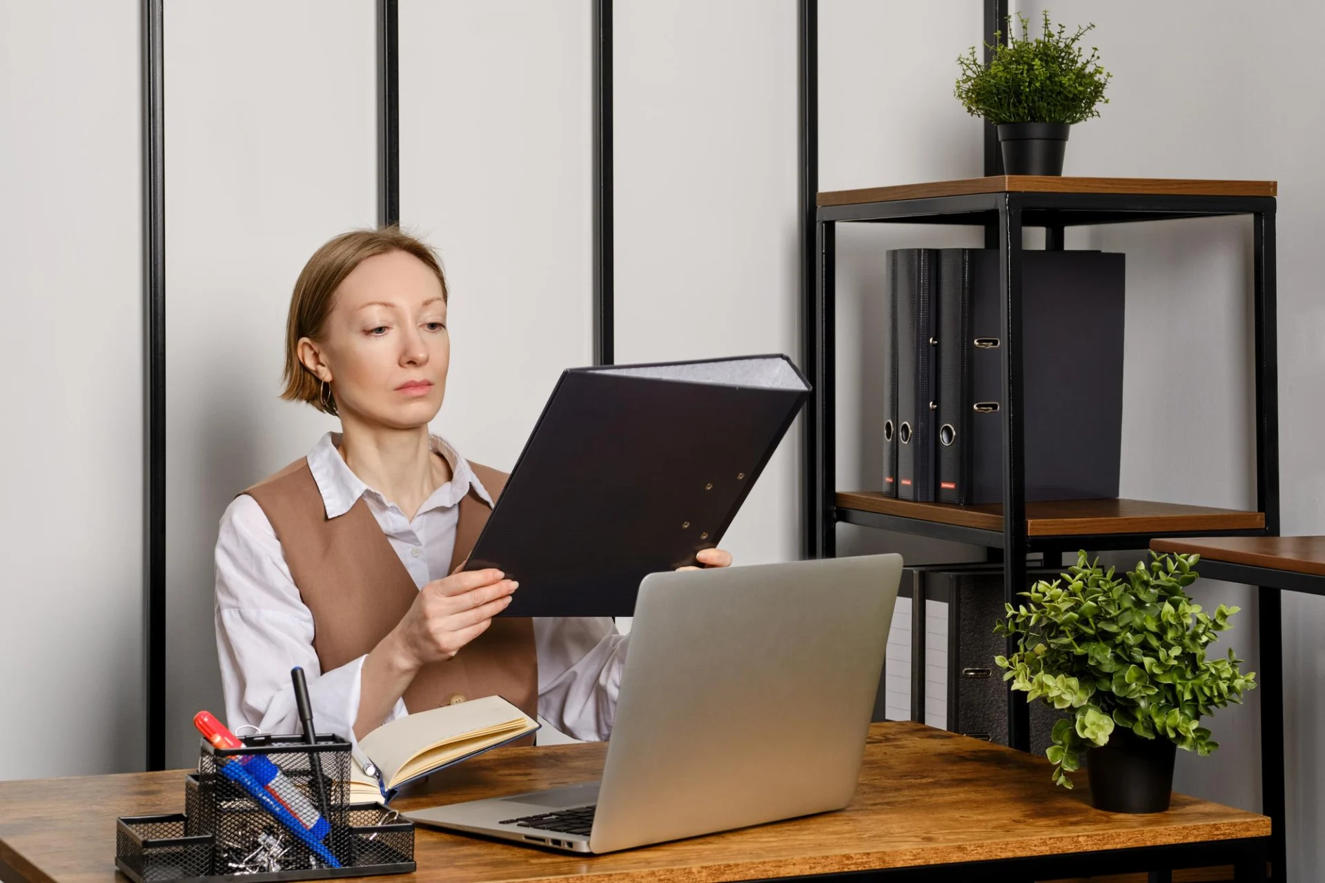 A quality professional at a desk examining a ring binder of assessment records, lever-arch files on the shelf behind