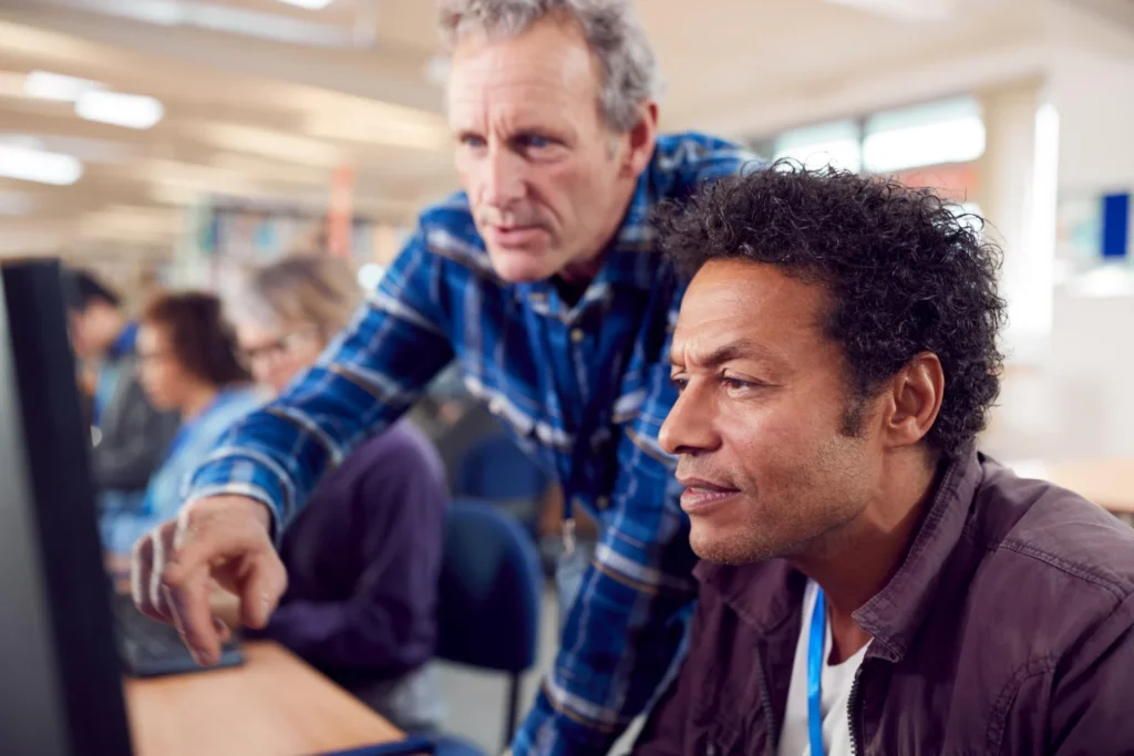 A tutor helping a mature adult learner at a computer in a further education college learning centre