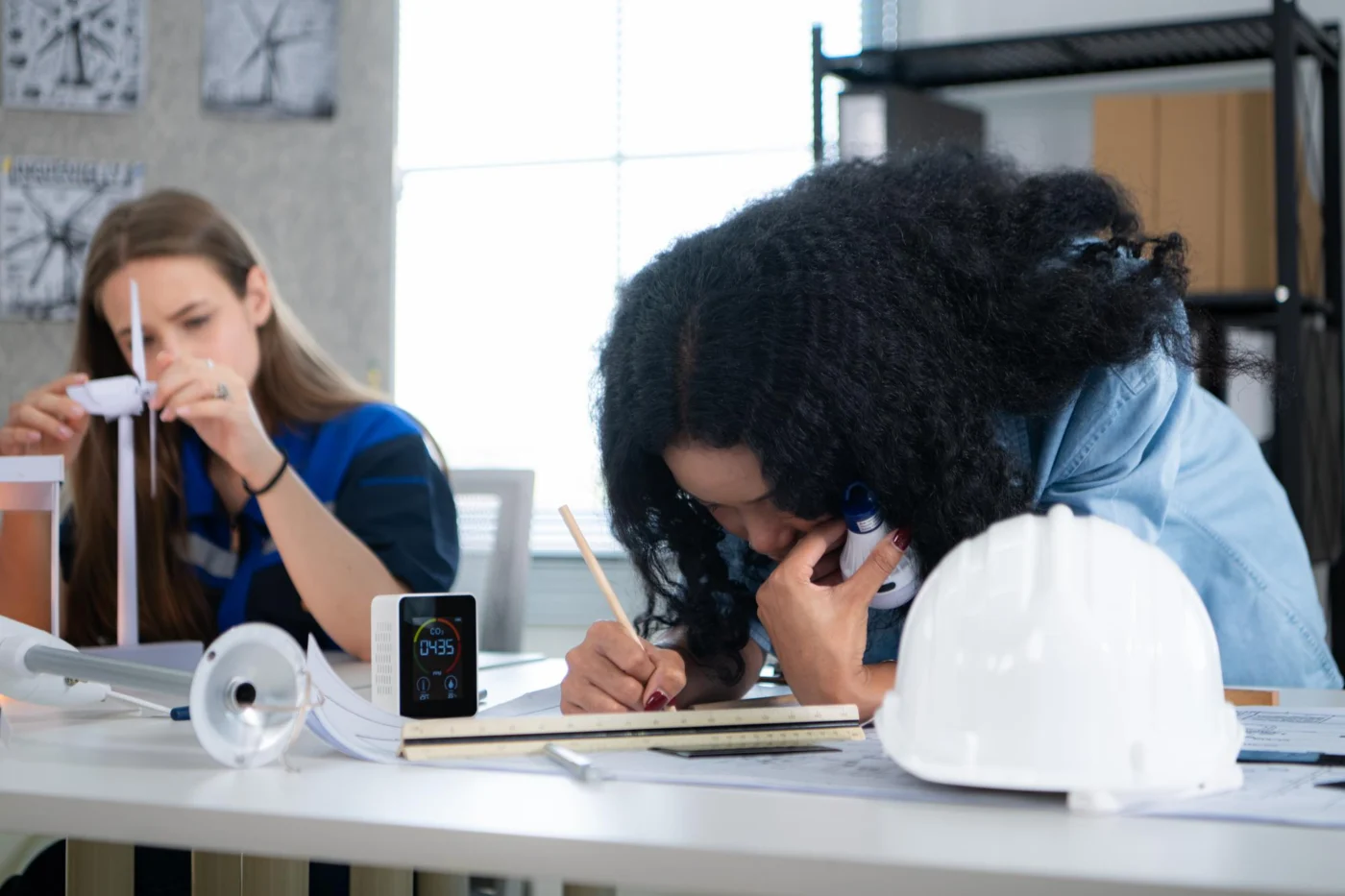 Two young women in an apprenticeship workshop wiring components and taking notes, one wearing a hard hat